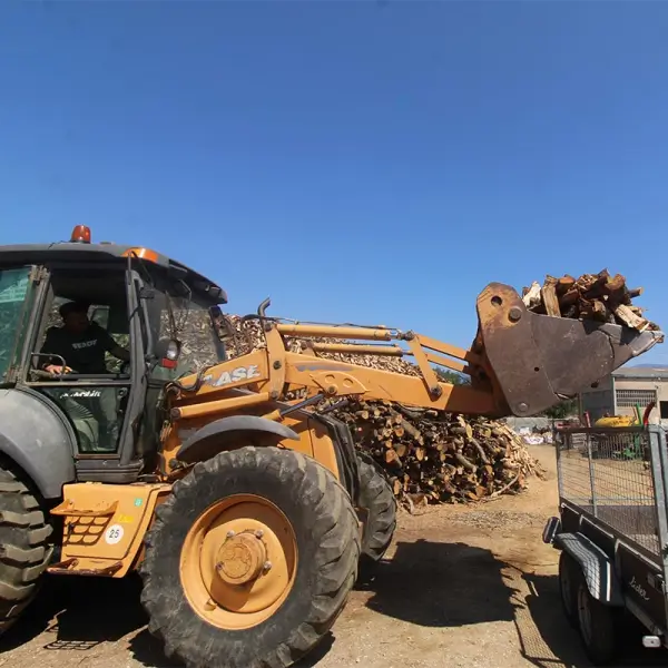 Tractor con pala cargando leña partida junto a un montón de leña y un remolque